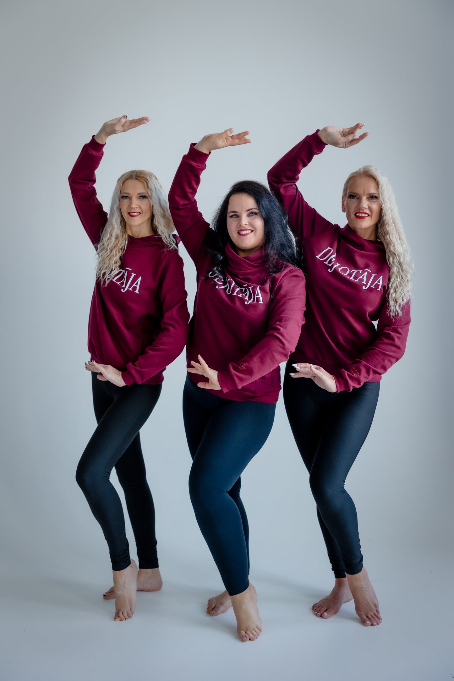 Three women wearing maroon sweatshirts with 'Dejotāja' printed on them, posing against a white background.