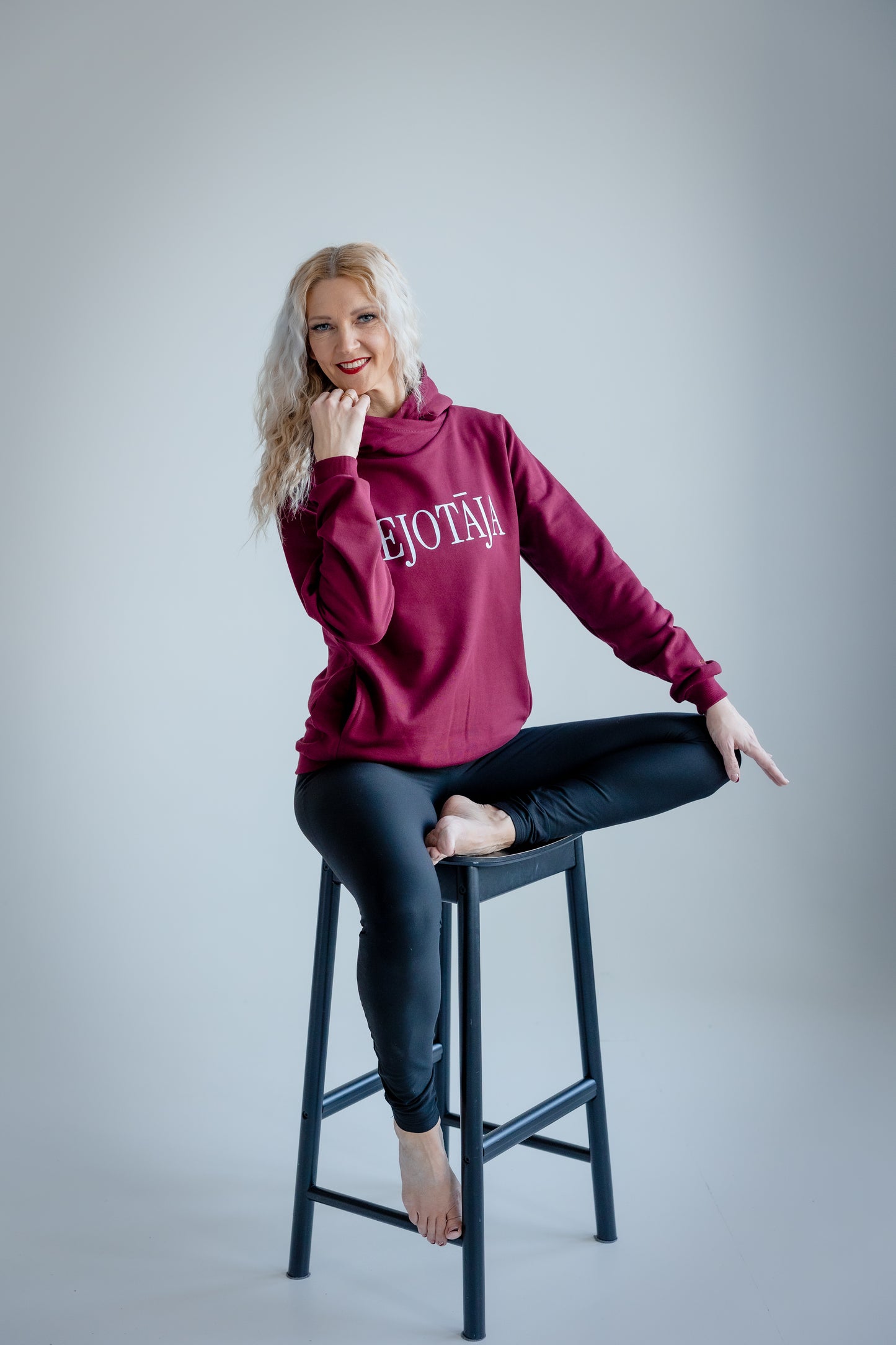 Woman wearing a maroon hoodie with text, sitting on a stool against a plain background