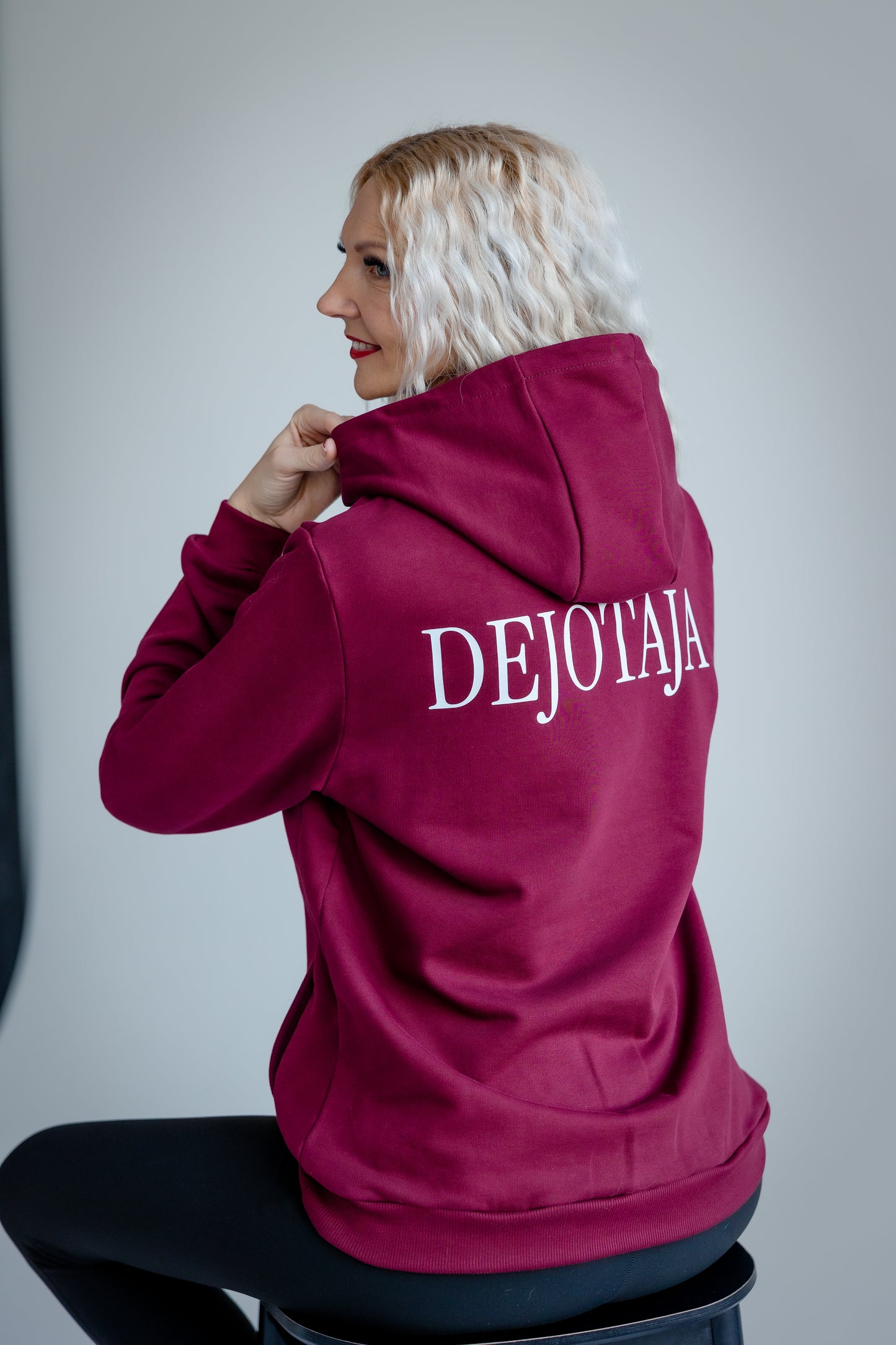 Person wearing a maroon hoodie with 'DEJOTĀJA' text on the back, sitting on a stool against a plain background.