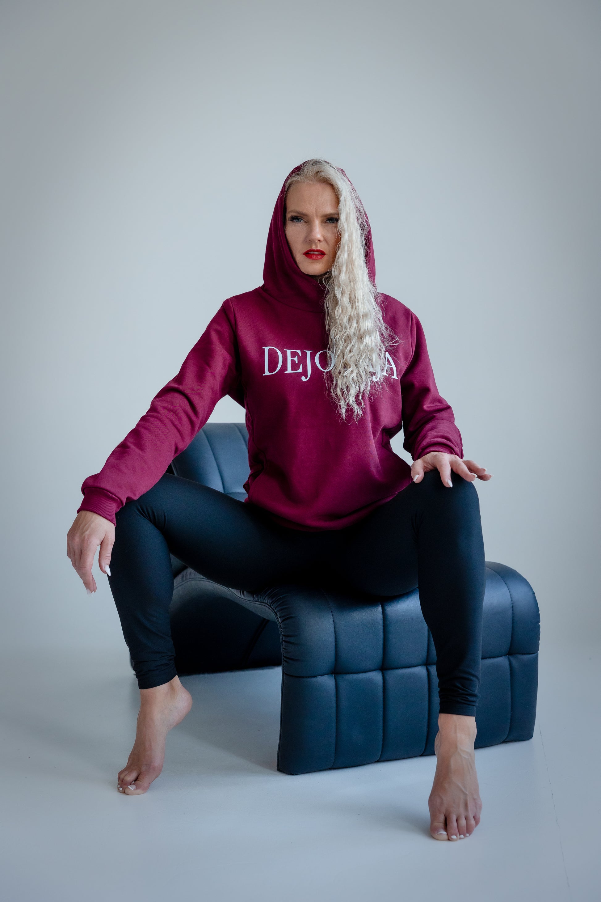 Person wearing a maroon hoodie with 'DEJOTĀJA' text, sitting on a blue chair against a gray background