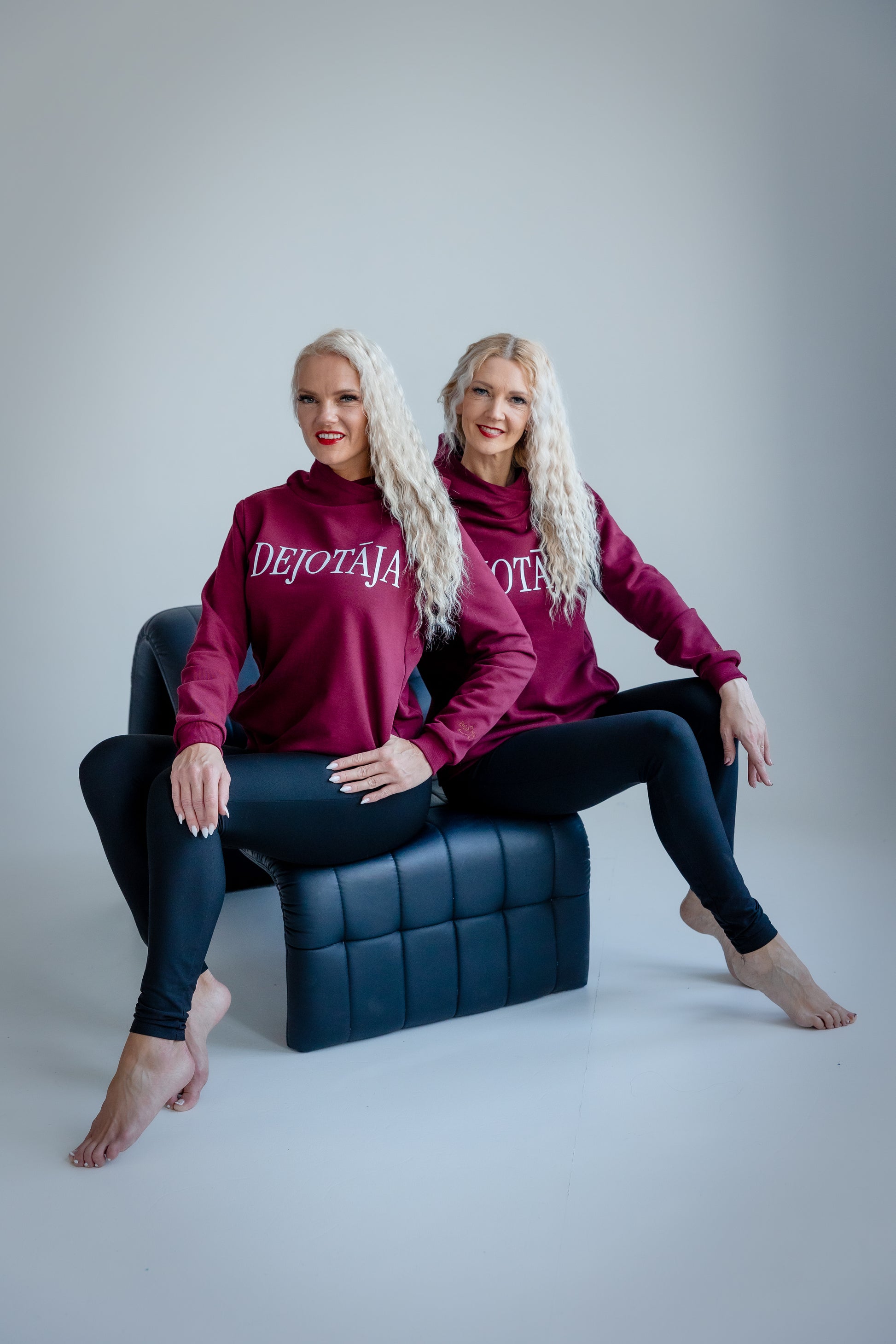 Two women wearing matching maroon hoodies with 'DEJOTAJA' text, sitting on a blue chair against a gray background.