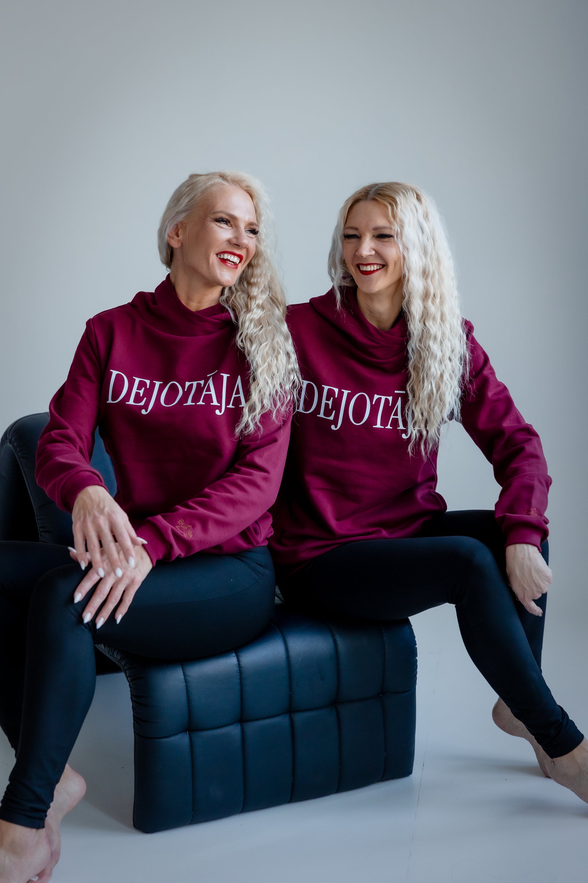 Two women wearing maroon sweatshirts with 'DEJOTAJA' text, sitting on a blue chair against a gray background.
