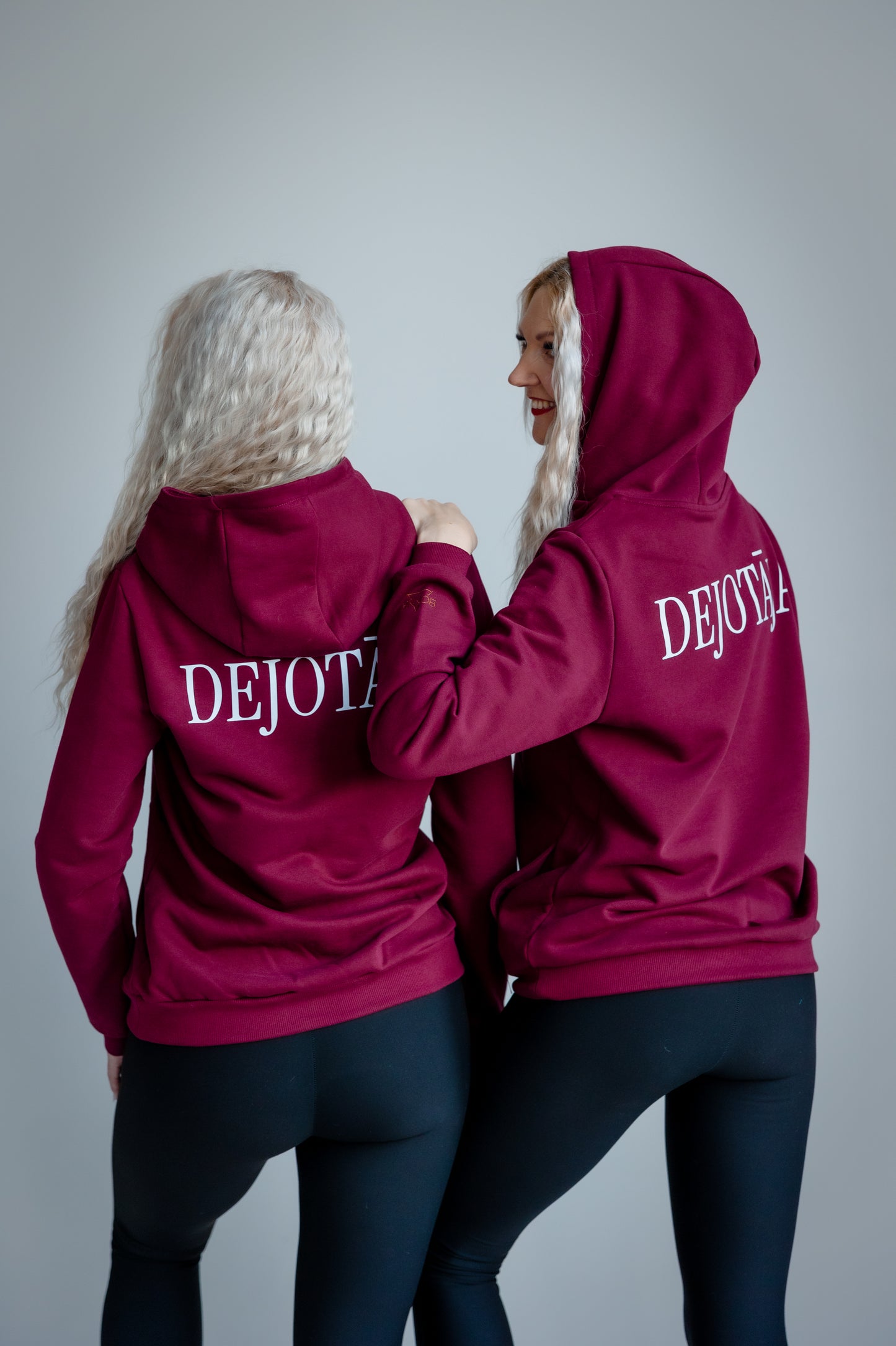 Two people wearing maroon hoodies with 'DEJOTĀJA' branding on a gray background