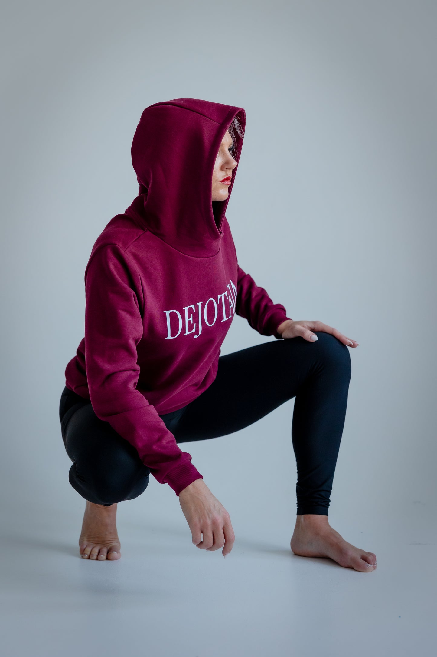 Person wearing a maroon hoodie with 'DEJOTĀJA' text, crouching on a light gray background