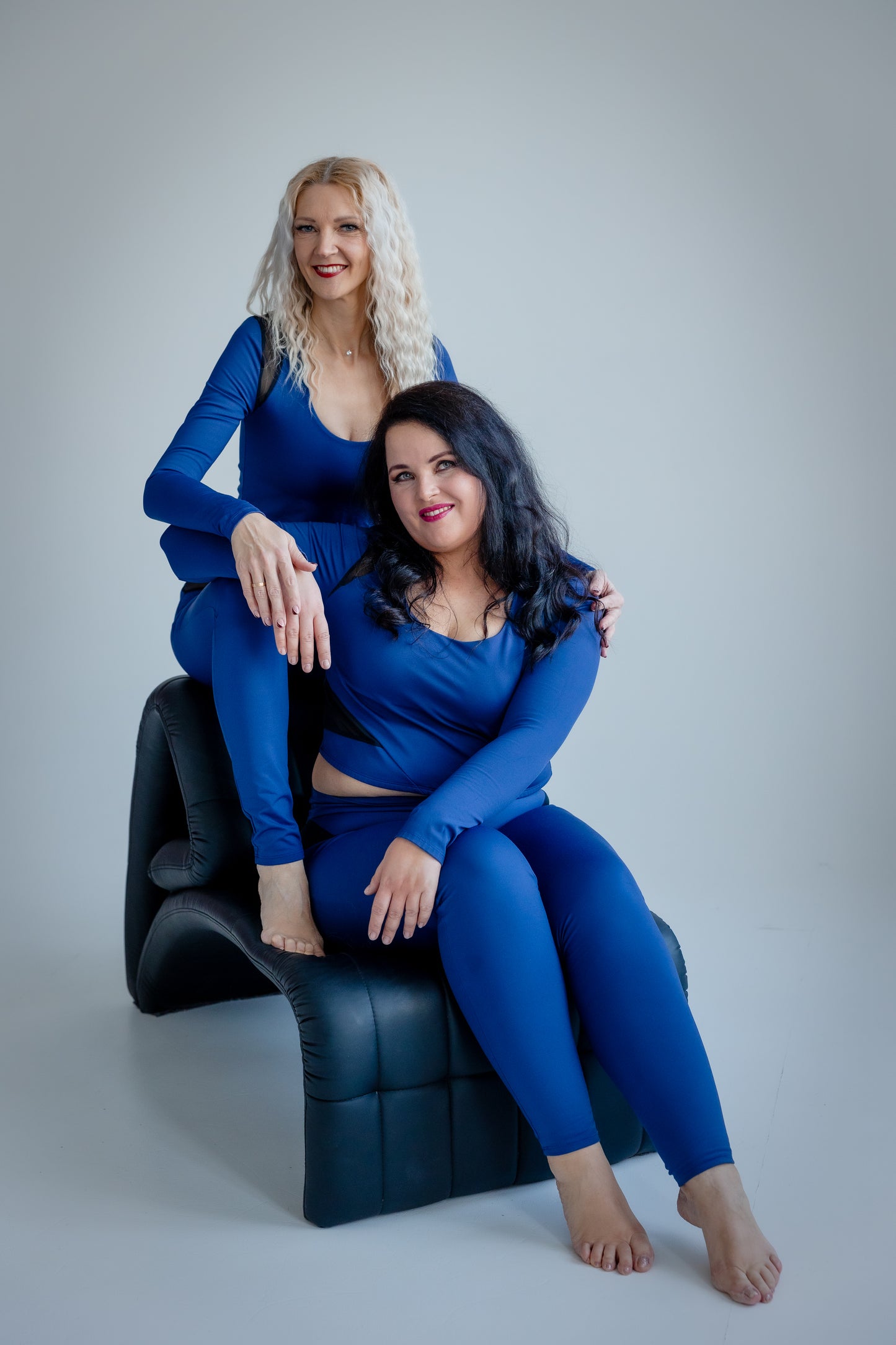 Two women in matching blue outfits sitting on a black chair against a gray background
