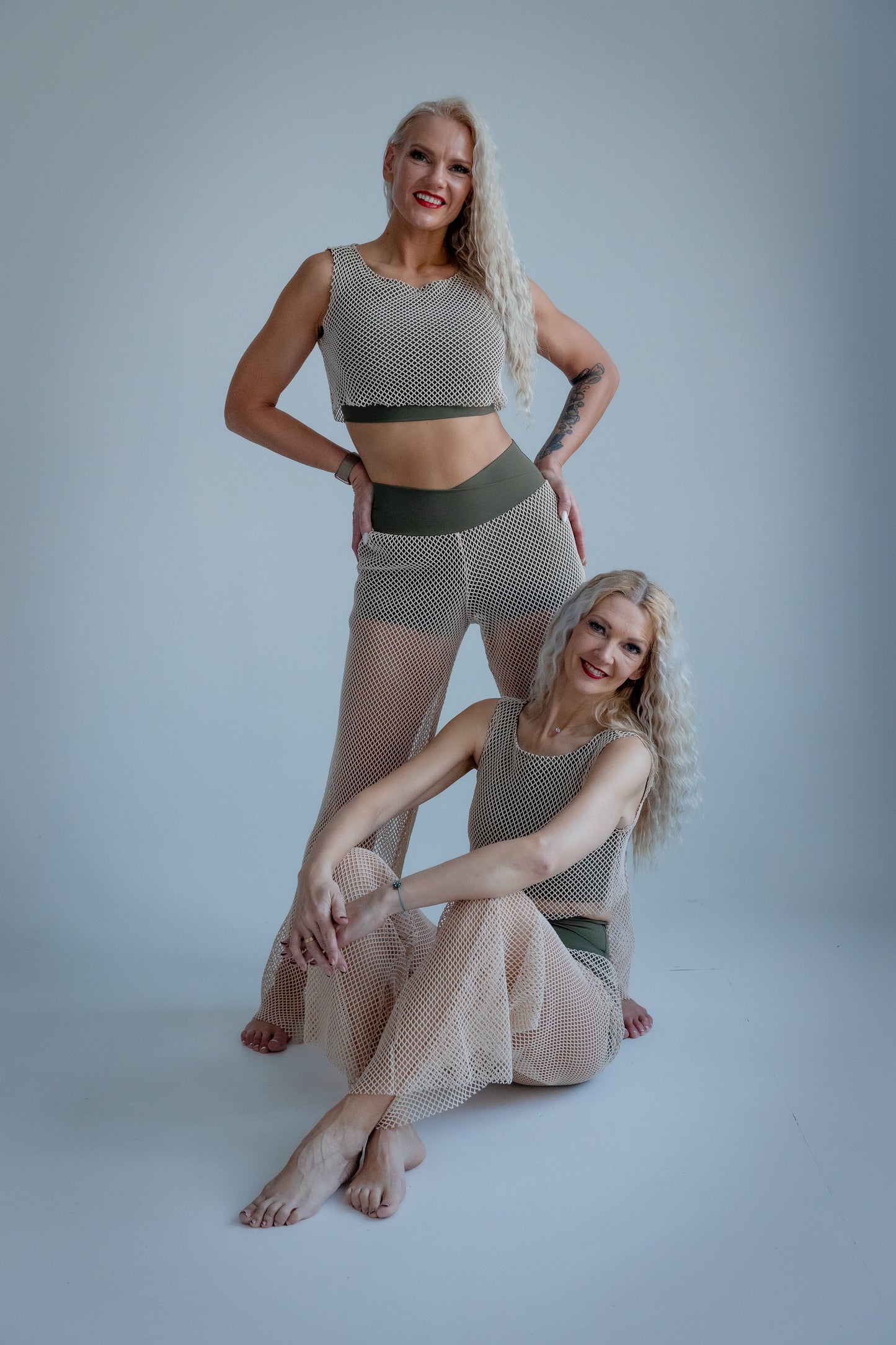 Two women wearing matching fishnet outfits on a plain background