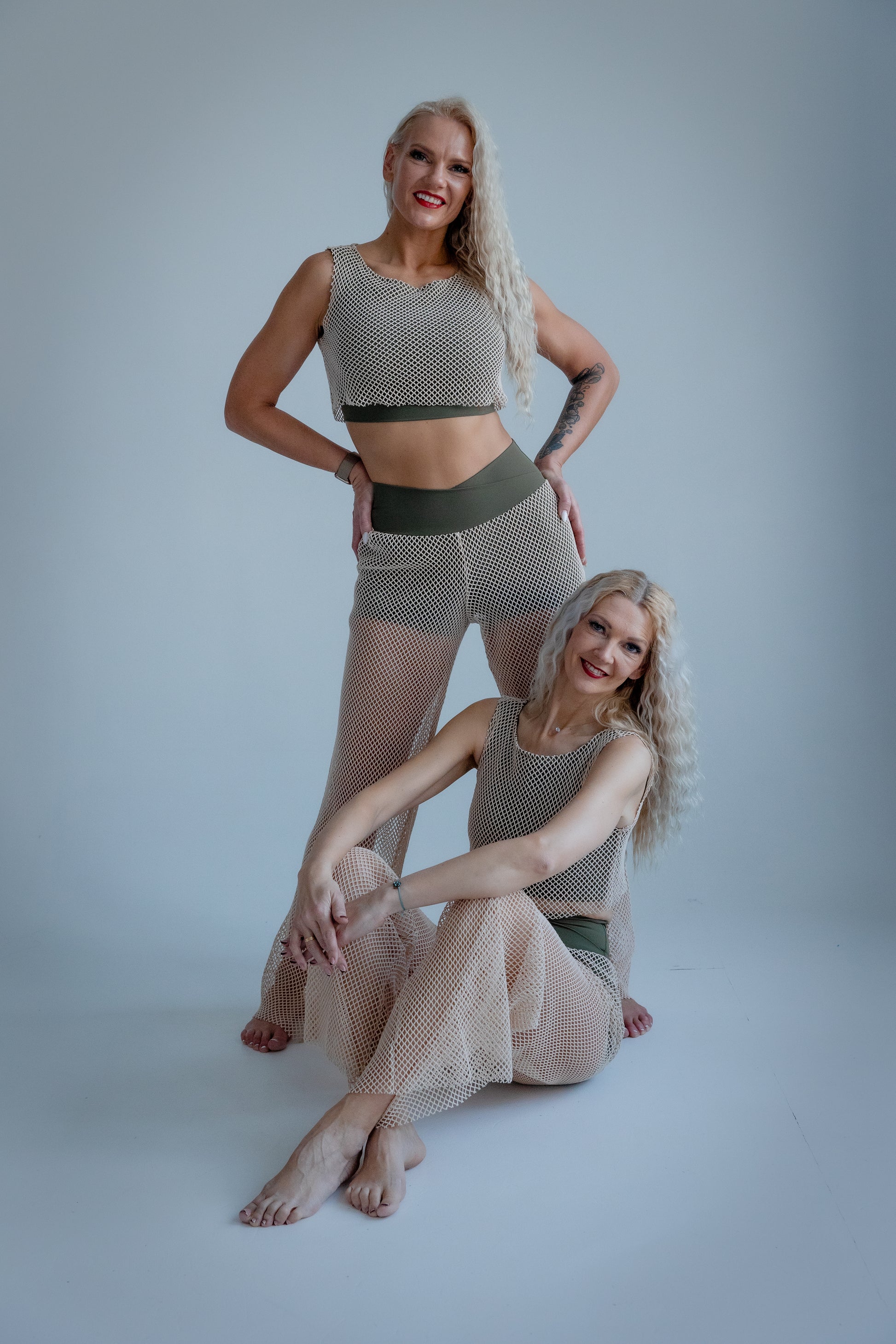 Two women wearing matching fishnet outfits on a plain background